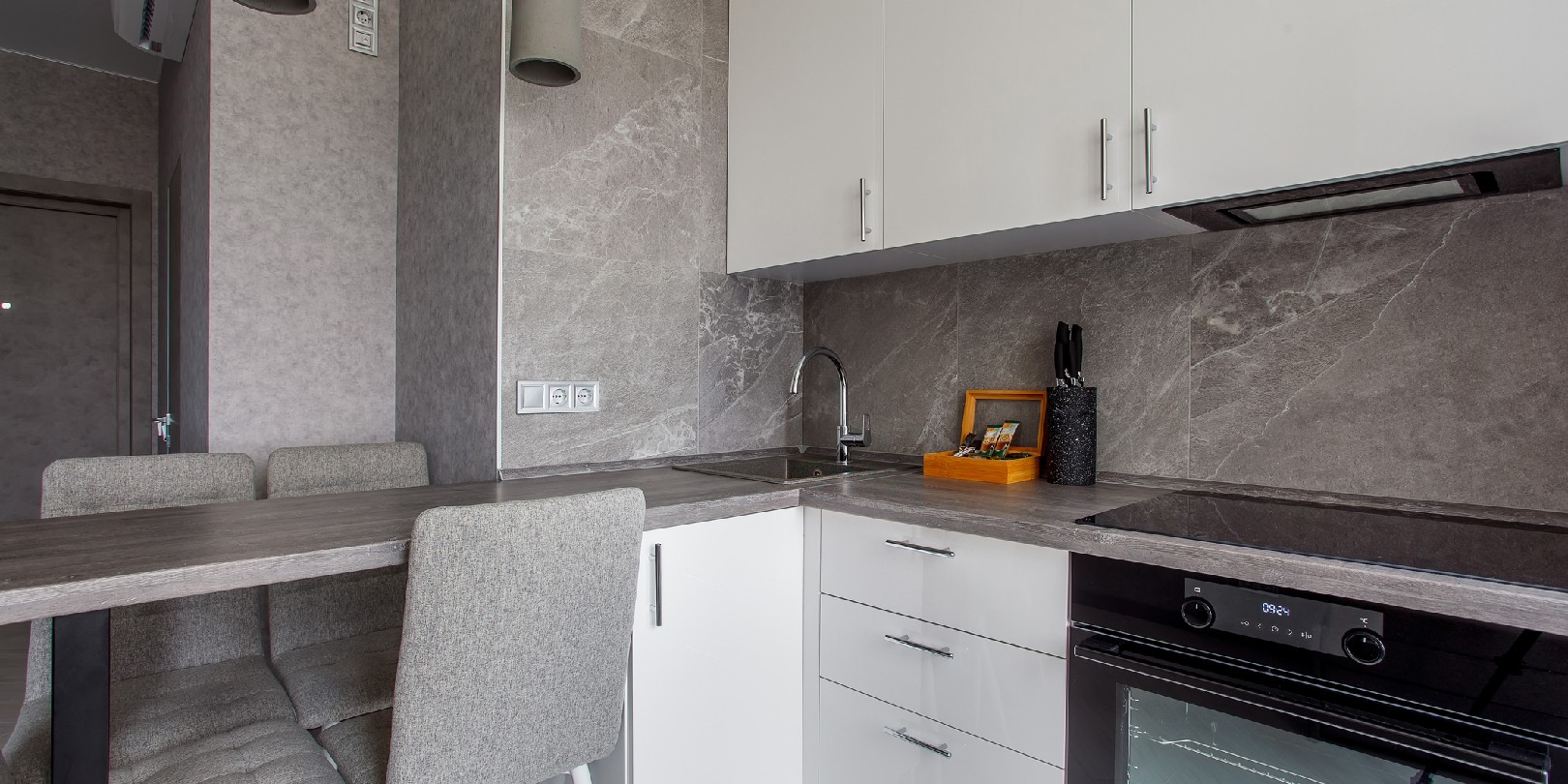 Cooking area by the window with lots of white and beige cabinets, countertop, sink, kitchen appliances by a tiled marble wall.